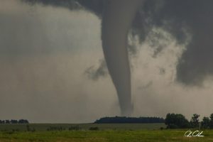 A large tornado funnel cloud above a flat landscape with trees and fields.