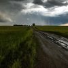 A stormy rural road stretching into the horizon, bordered by green fields and under a looming, dramatic sky.